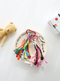 Two colorful rope keychains with multiple knotted strands — one in pink, black, and white tones, the other in bright rainbow colors — each with playful bead details, small smiley charms, and metal clasps for easy attachment on a white text written plate.