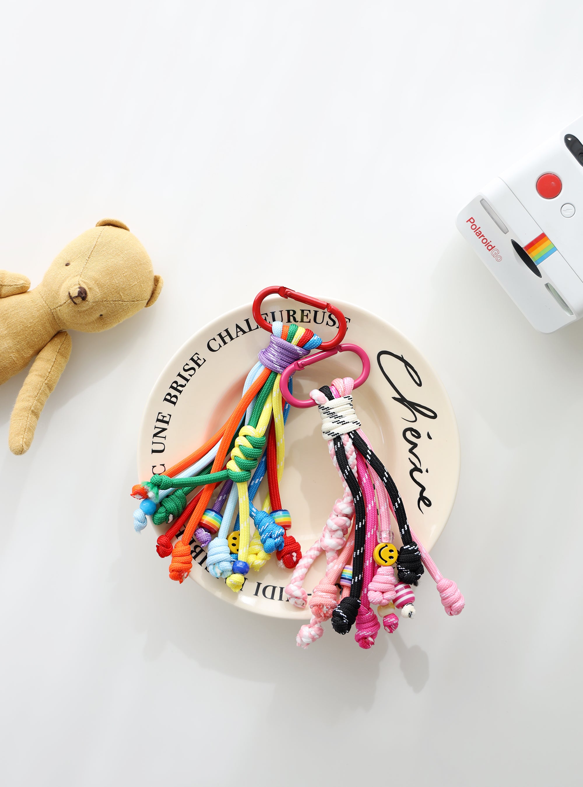 Two colorful rope keychains with multiple knotted strands — one in pink, black, and white tones, the other in bright rainbow colors — each with playful bead details, small smiley charms, and metal clasps for easy attachment on a white text written plate.