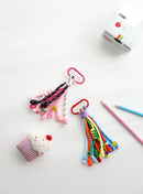Two playful rope keychains with knotted strands — one in pink, black, and white tones, and one in bright rainbow colors — each with bead details, smiley charms, and sturdy clasps, displayed on a white background with a Polaroid camera, pencils, and a felt cupcake for a fun, colorful touch.