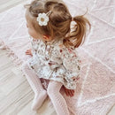 Little girl sitting on a light pink rug, wearing a floral dress and pale pink knit tights, with a cream crochet flower hair clip on a silver snap clip in her side ponytail.
