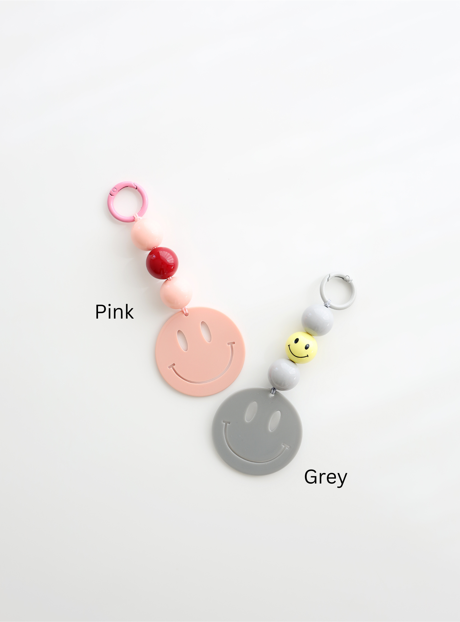 Two colorful smiley face keychains, one in pink and one in grey, each featuring round pastel beads and a large smiley face charm, labeled with their color names on a white background.