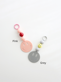 Two colorful smiley face keychains, one in pink and one in grey, each featuring round pastel beads and a large smiley face charm, labeled with their color names on a white background.