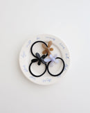 Three black elastic hair ties, each featuring a glossy flower-shaped charm in beige, lavender, and black, displayed on a white plate with handwritten text.