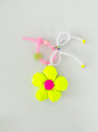 Neon yellow five-petal flower bag charm with a pink center, pearl bead bow, and pink silicone clasp. Clasp has small clips in various shapes and colors, shown here as a green flower and pink crown.