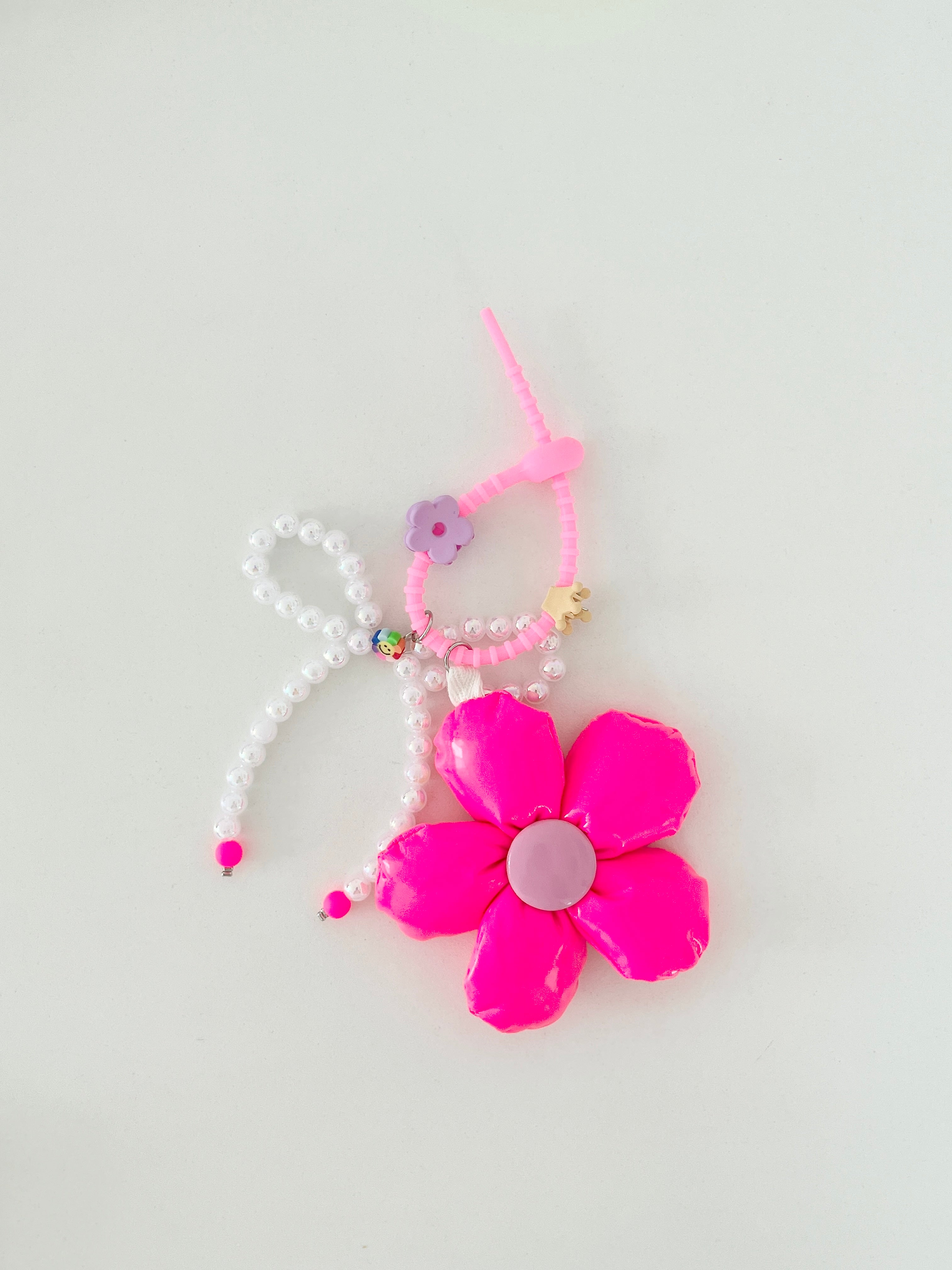 Neon pink five-petal flower bag charm with a light pink center, pearl bead bow, and pink silicone clasp. Clasp has small clips in various shapes and colors, shown here as a purple flower and beige crown.
