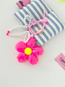Neon pink five-petal flower bag charm with a yellow center, pearl bead bow, and pink silicone clasp. Clasp has small clips in various shapes and colors, shown here as a purple flower and gold crown.
