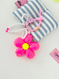 Neon pink five-petal flower bag charm with a yellow center, pearl bead bow, and pink silicone clasp. Clasp has small clips in various shapes and colors, shown here as a purple flower and gold crown.