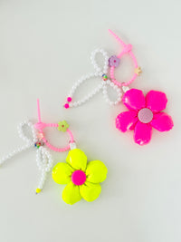 Two bag charms with pearl bead bows and silicone clasps. One has a neon pink flower with a light pink center, and the other has a neon yellow flower with a pink center. Clasp clips vary in shape and color, shown here as a purple flower and a green flower.