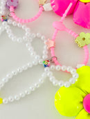 Close-up of two bag charms showing pearl bead bows and pink silicone clasps. One clasp has a purple flower clip and a small multicolor smiley charm, and the other has a green flower clip and a small gold crown clip. Part of a neon pink flower and a neon yellow flower are visible.