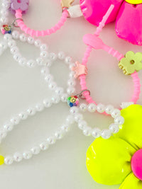 Close-up of two bag charms showing pearl bead bows and pink silicone clasps. One clasp has a purple flower clip and a small multicolor smiley charm, and the other has a green flower clip and a small gold crown clip. Part of a neon pink flower and a neon yellow flower are visible.