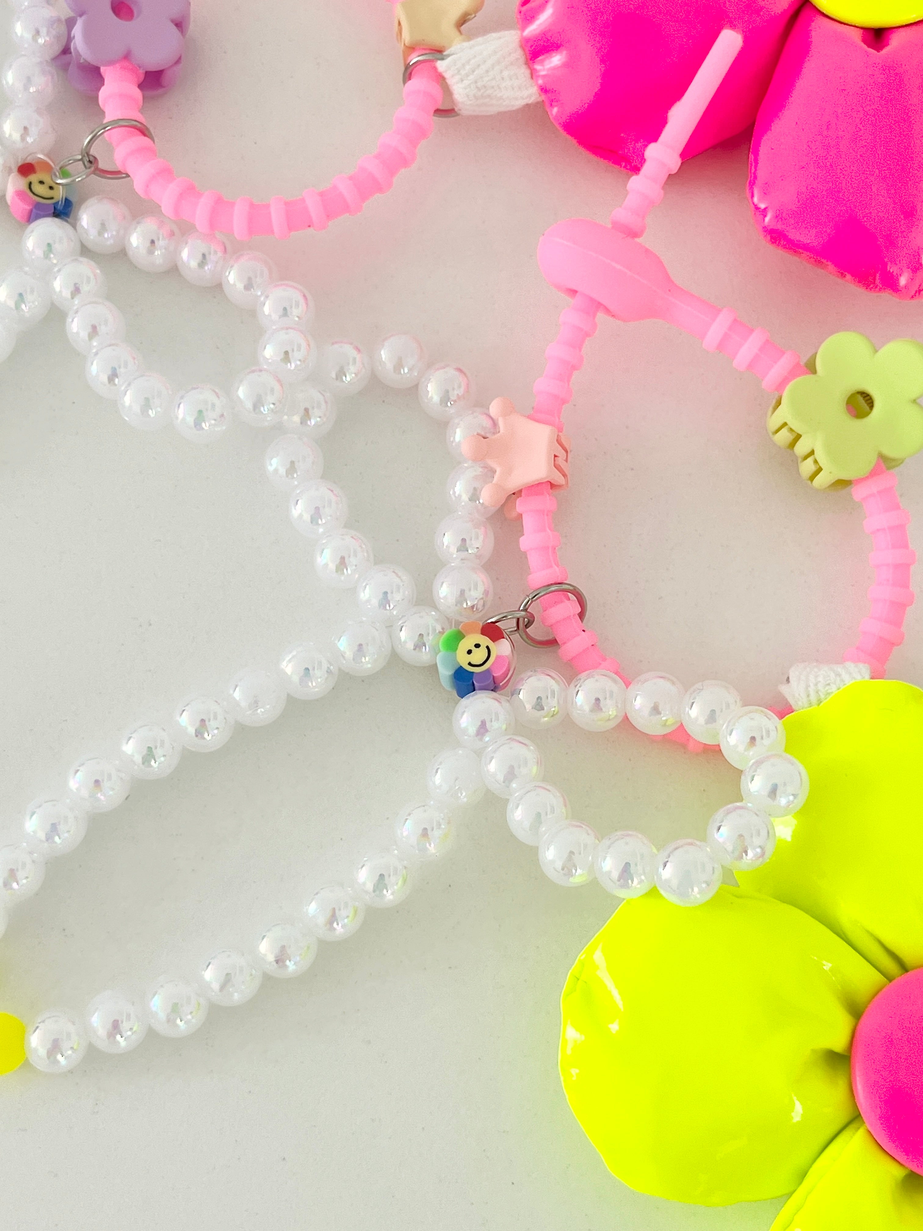 Close-up of two bag charms showing pearl bead bows and pink silicone clasps. One clasp has a purple flower clip and a small multicolor smiley charm, and the other has a green flower clip and a small gold crown clip. Part of a neon pink flower and a neon yellow flower are visible.