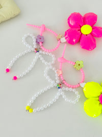 Two bag charms with pearl bead bows and silicone clasps. One has a neon pink flower with a yellow center, and the other has a neon yellow flower with a pink center. Clasp clips vary in shape and color, shown here as a purple flower and a green flower.