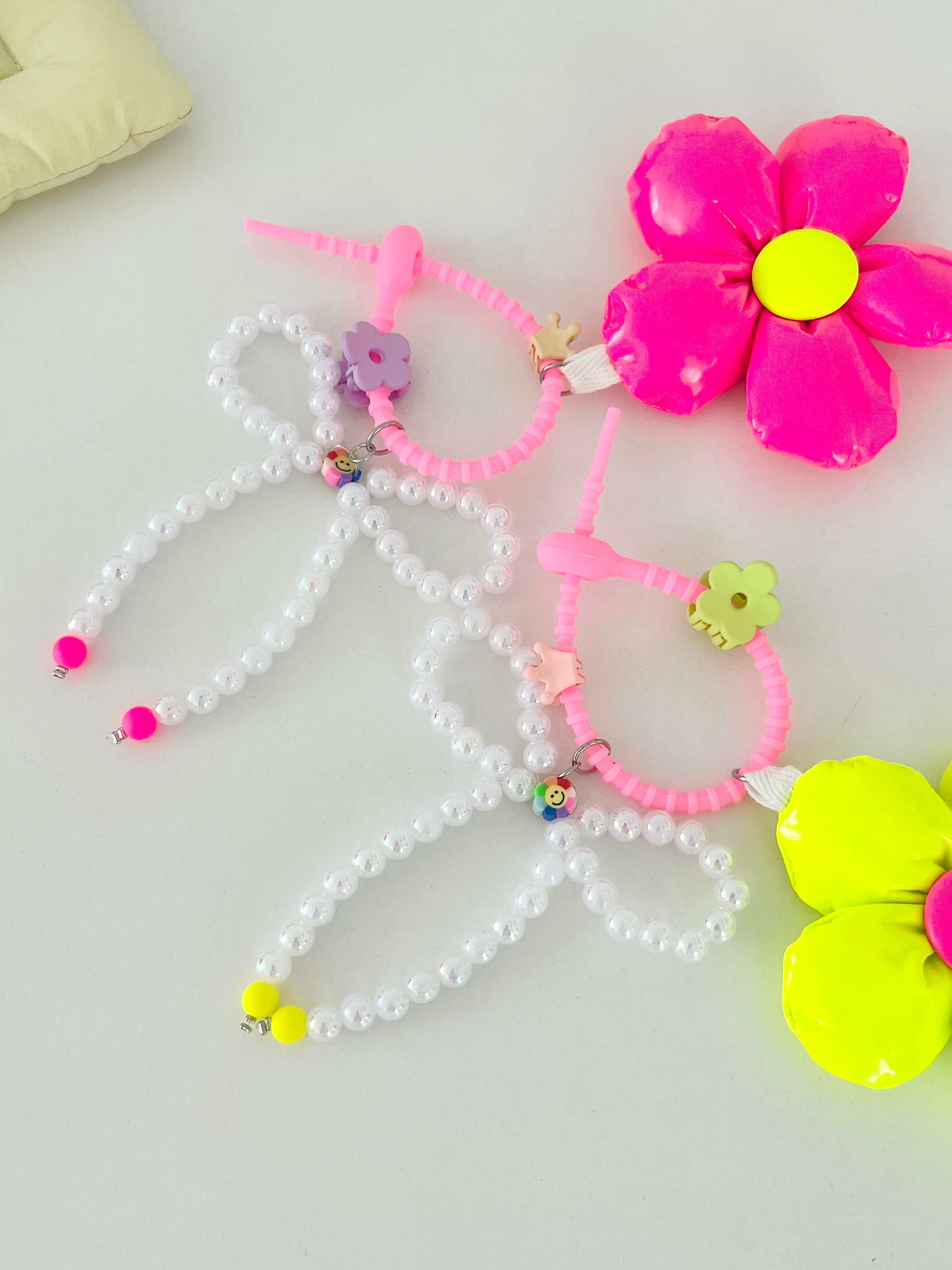Two bag charms with pearl bead bows and silicone clasps. One has a neon pink flower with a yellow center, and the other has a neon yellow flower with a pink center. Clasp clips vary in shape and color, shown here as a purple flower and a green flower.