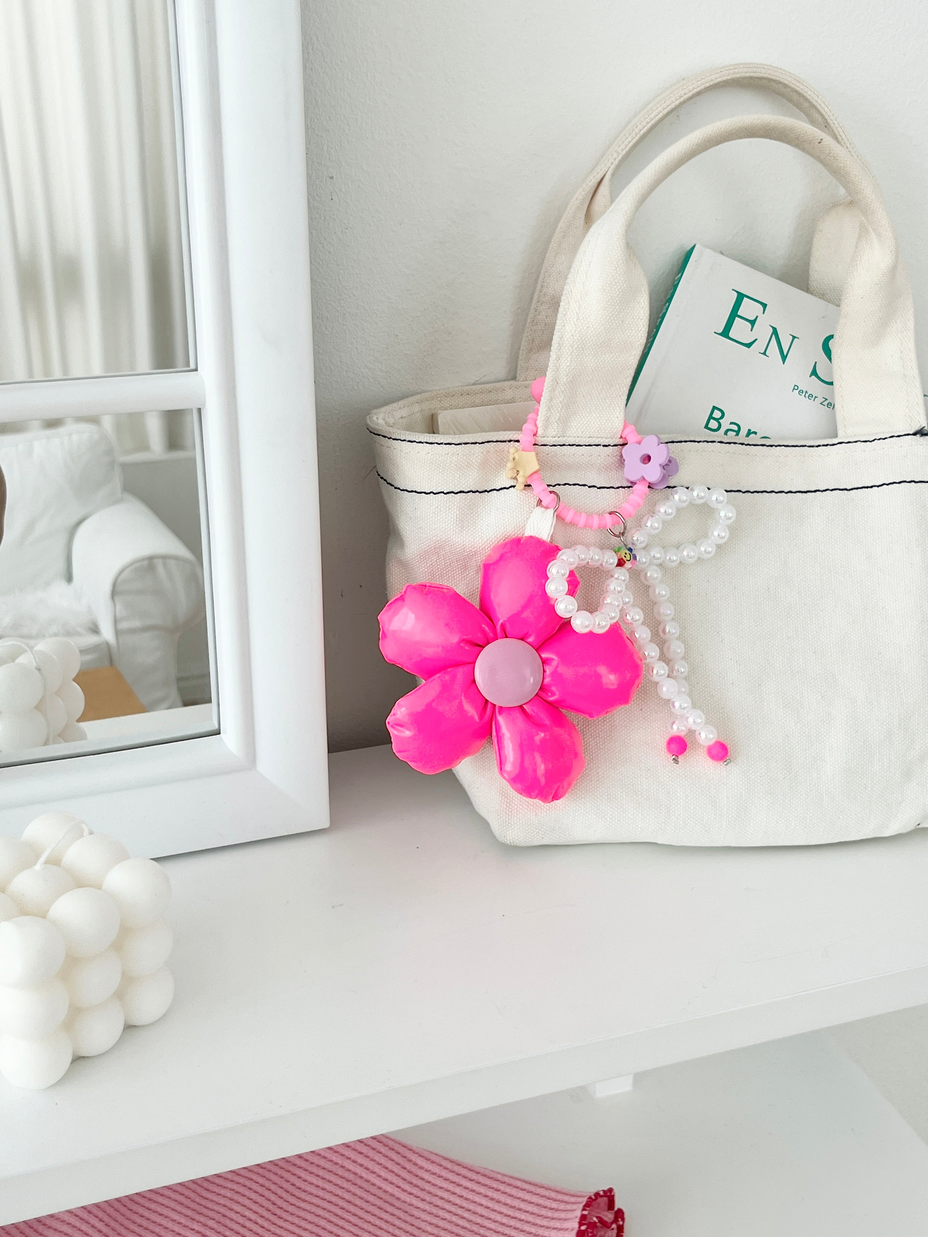 Canvas tote bag with a neon pink five-petal flower charm featuring a light pink center, pearl bead bow, and pink silicone clasp. Clasp clips shown as a purple flower and gold crown, with a small multicolor smiley charm.