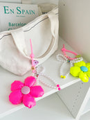 Two bag charms with pearl bead bows and silicone clasps. One has a neon pink flower with a light purple center, and the other has a neon yellow flower with a blue center. Clasp clips vary in shape and color, shown here as a purple flower and a green flower.