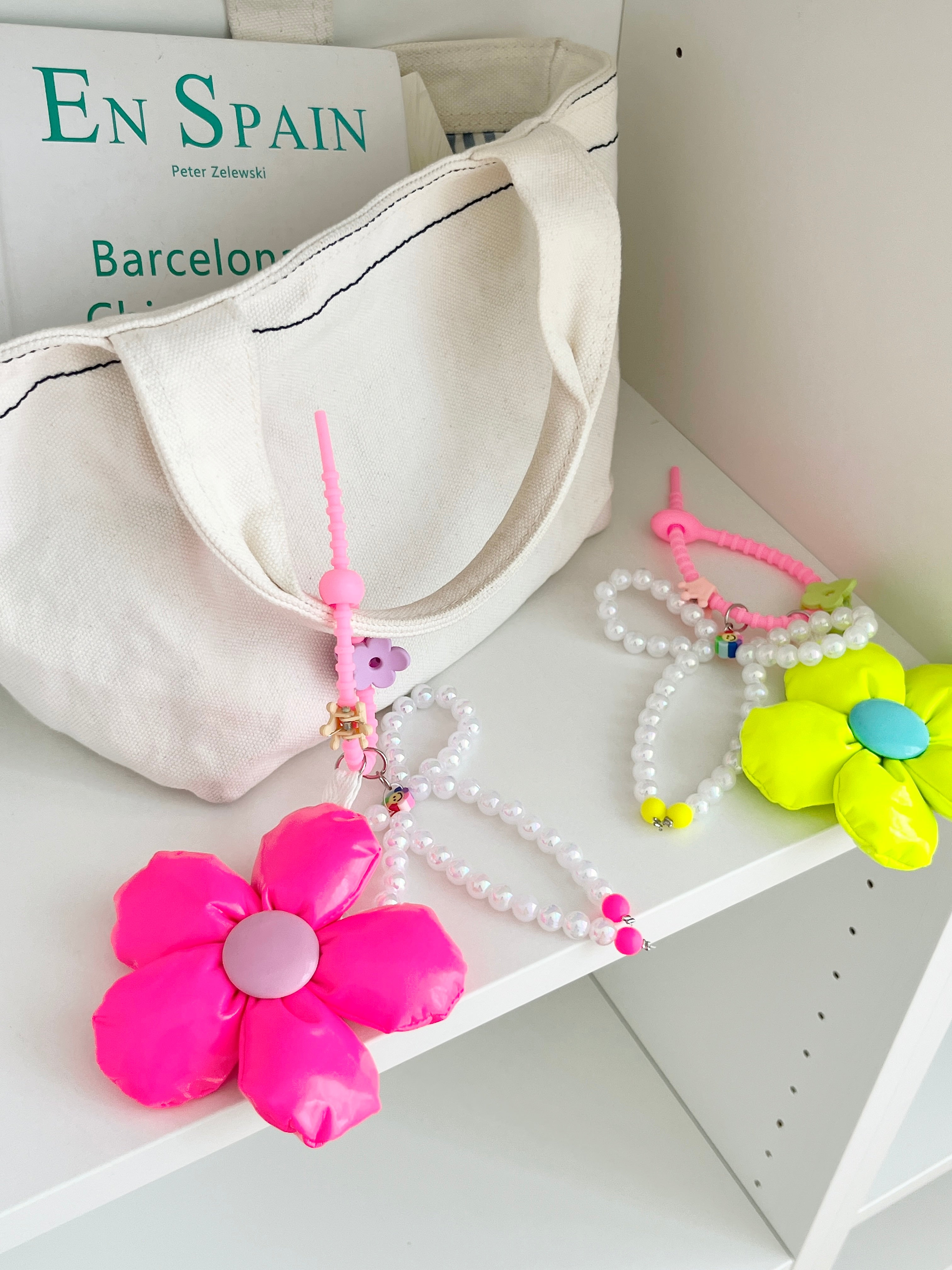 Two bag charms with pearl bead bows and silicone clasps. One has a neon pink flower with a light purple center, and the other has a neon yellow flower with a blue center. Clasp clips vary in shape and color, shown here as a purple flower and a green flower.