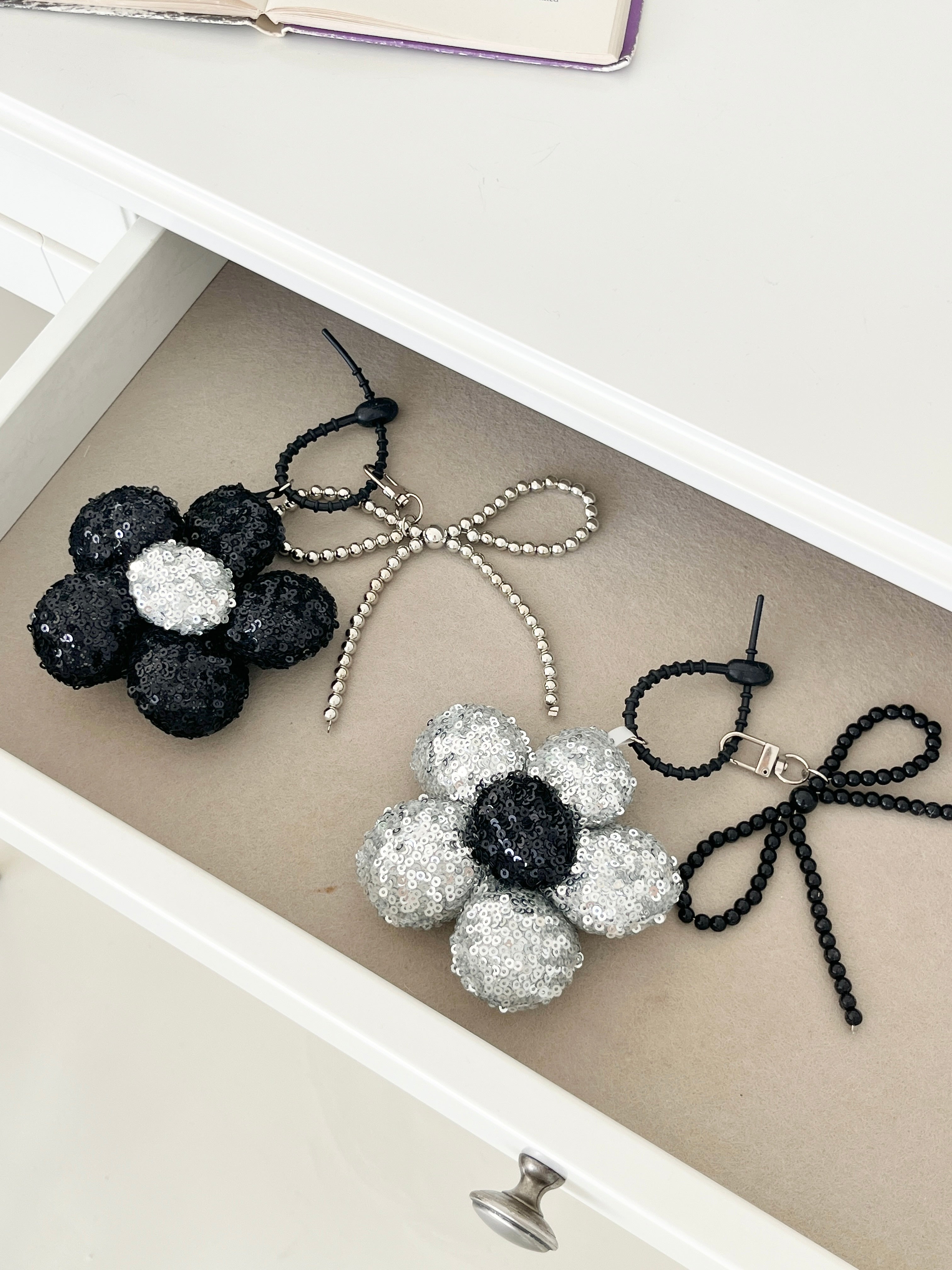 Two sequined flower keychains in black and silver with matching bead bow charms, neatly arranged in a drawer next to each other.