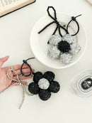 Two sequined flower keychains, one black with a silver center and one silver with a black center, shown one on a white plate and the other one on a hand.