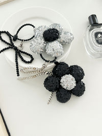 Two sequined flower keychains in black and silver with matching bead bow charms, displayed on a white plates next to perfume bottle.