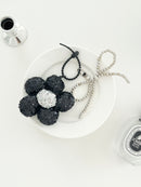 A black sequined flower keychains with silver center and matching bead bow charms, displayed on a white plates next to perfume bottle.