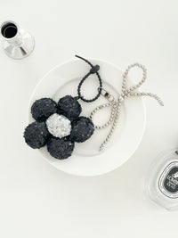 A black sequined flower keychains with silver center and matching bead bow charms, displayed on a white plates next to perfume bottle.