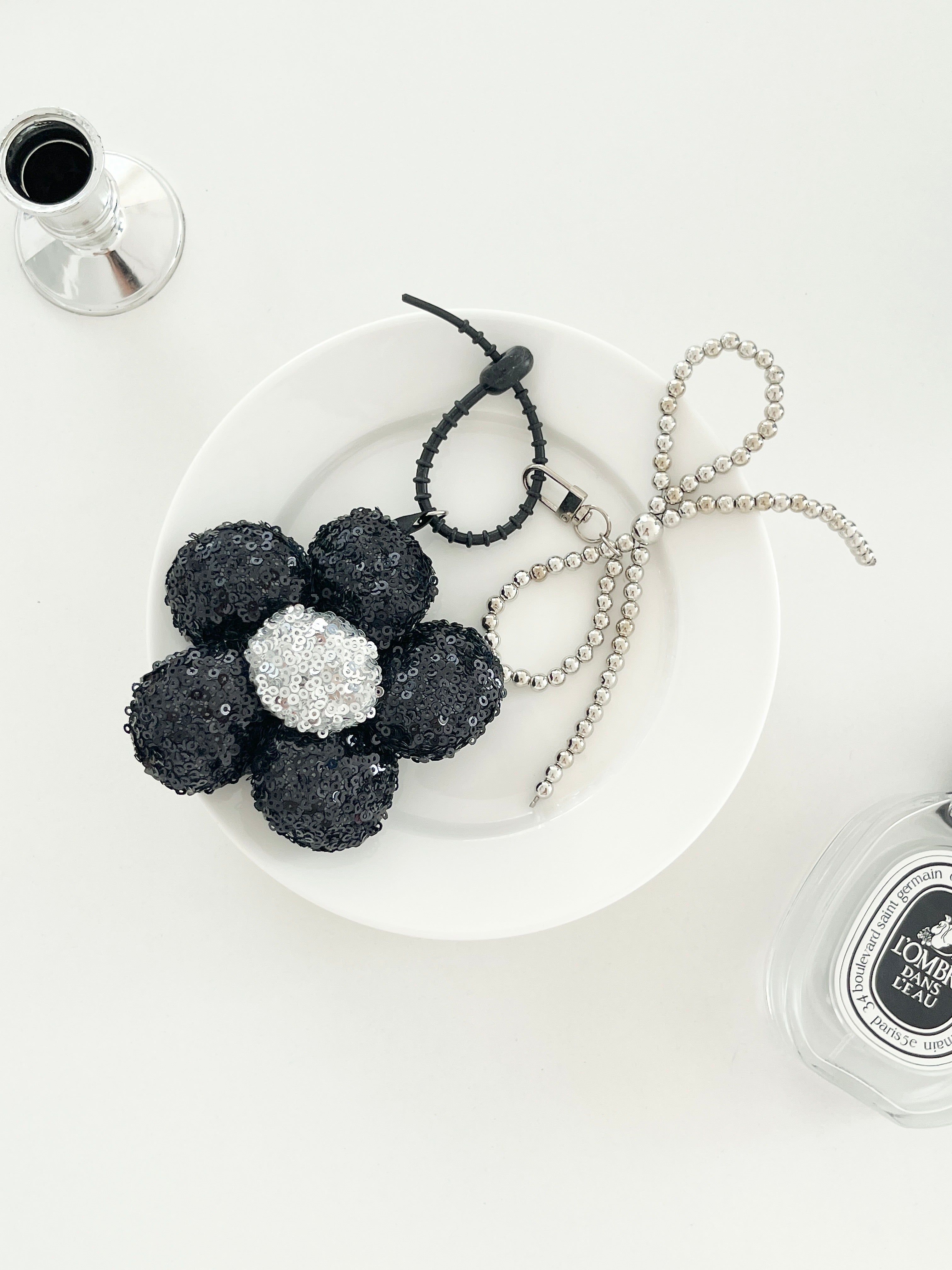 A black sequined flower keychains with silver center and matching bead bow charms, displayed on a white plates next to perfume bottle.