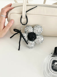A hand holds the strap of a cream handbag with a silver sequined flower keychain featuring a black center and beaded bow attached.