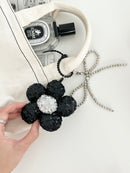 A hand holds a black sequined flower keychain with a silver center, attached to a cream tote bag with a perfume bottle inside.