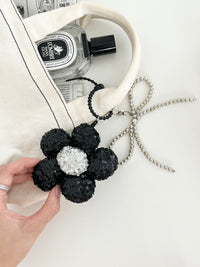 A hand holds a black sequined flower keychain with a silver center, attached to a cream tote bag with a perfume bottle inside.