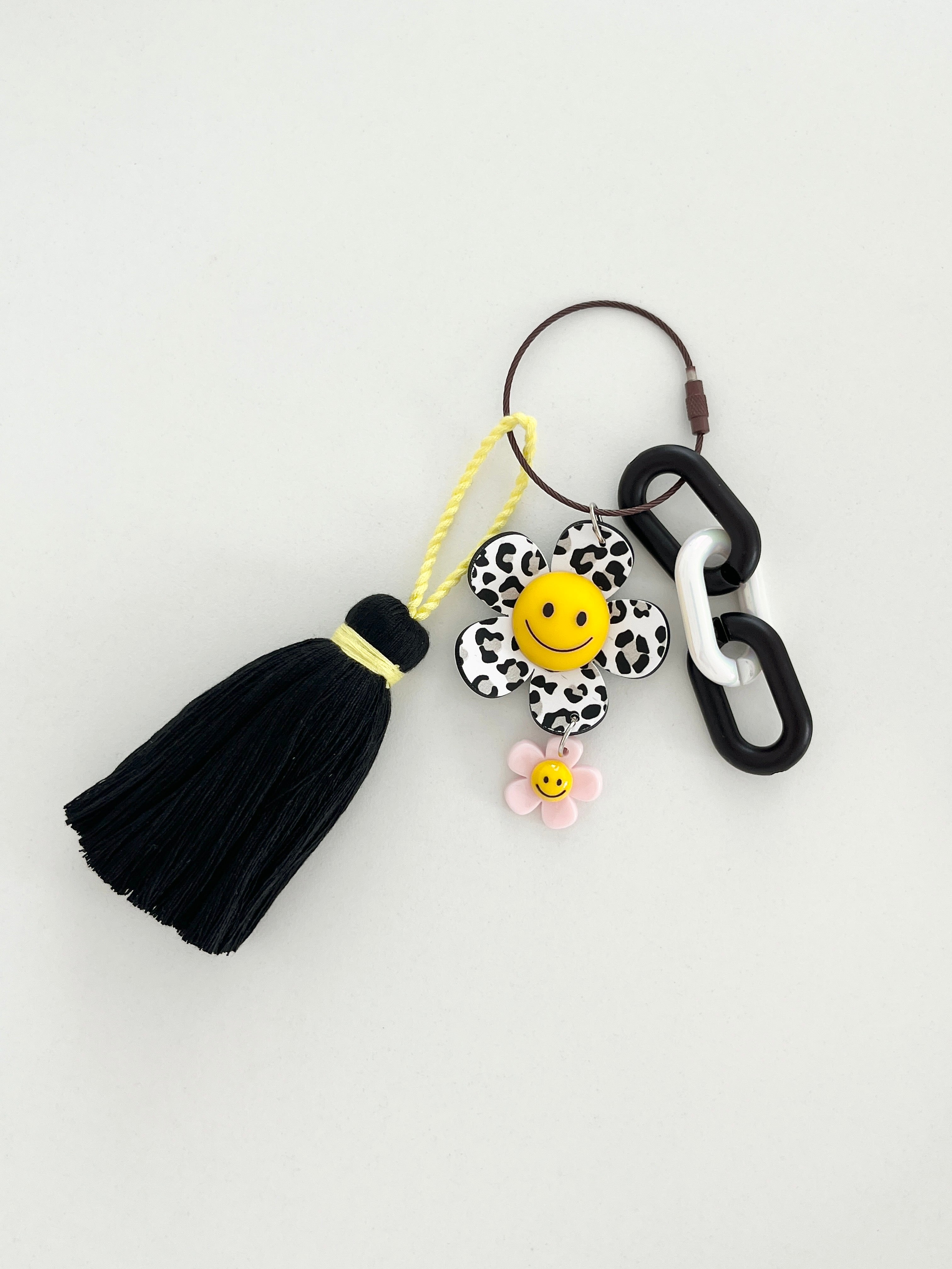 Black and white leopard print flower keychain with a yellow smiley face center, small yellow flower charm, and a black tassel hanging from chunky black and white links displayed in white background. 