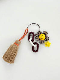 Leopard print flower keychain with a yellow smiley face center, small yellow flower charm, and a tan tassel hanging from chunky dark brown and white links displayed in white background. 