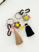 Two decorative keychains with smiley face flowers in animal print designs — one in brown leopard print with a tan tassel and one in black-and-white zebra print with a black tassel. Both have chunky linked rings and small accent charms attached, displayed on a light background.