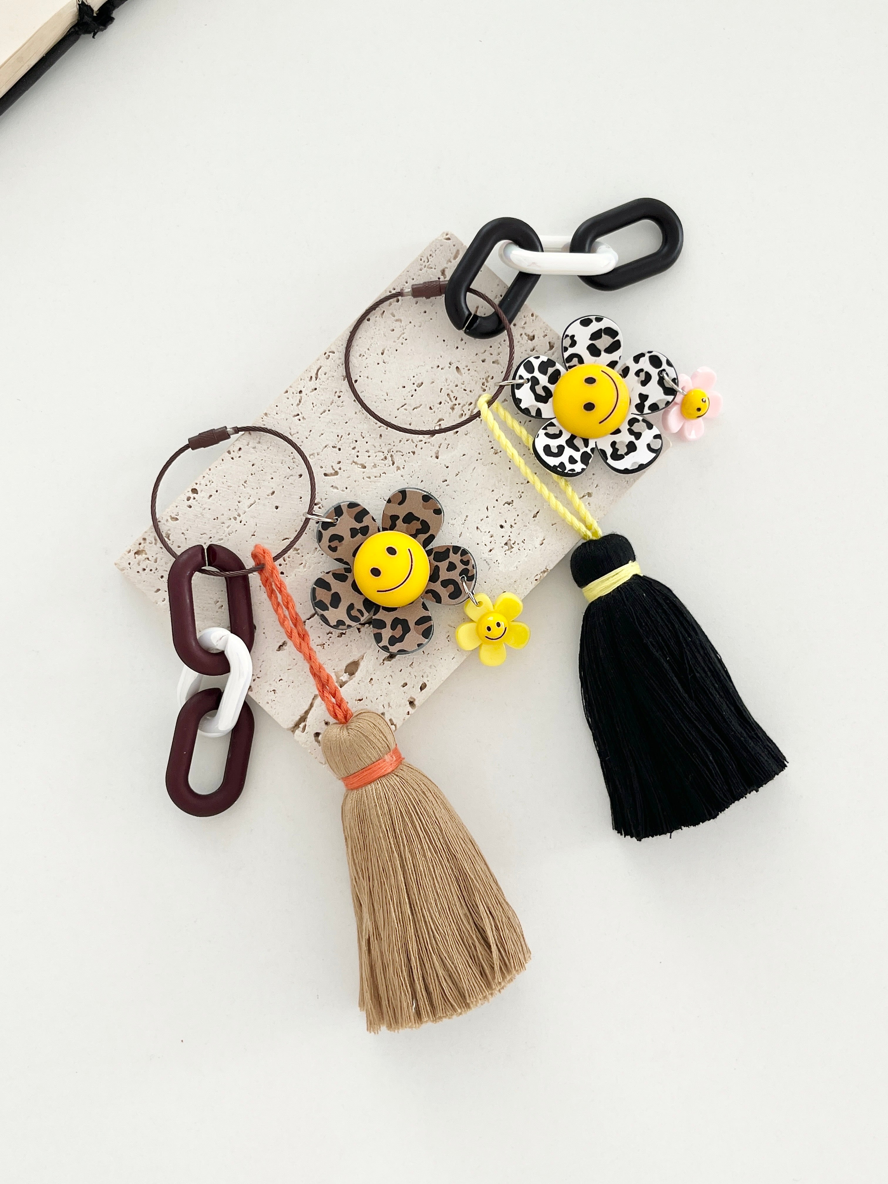 Two decorative keychains with smiley face flowers in animal print designs — one in brown leopard print with a tan tassel and one in black-and-white zebra print with a black tassel. Both have chunky linked rings and small accent charms attached, displayed on a light background.