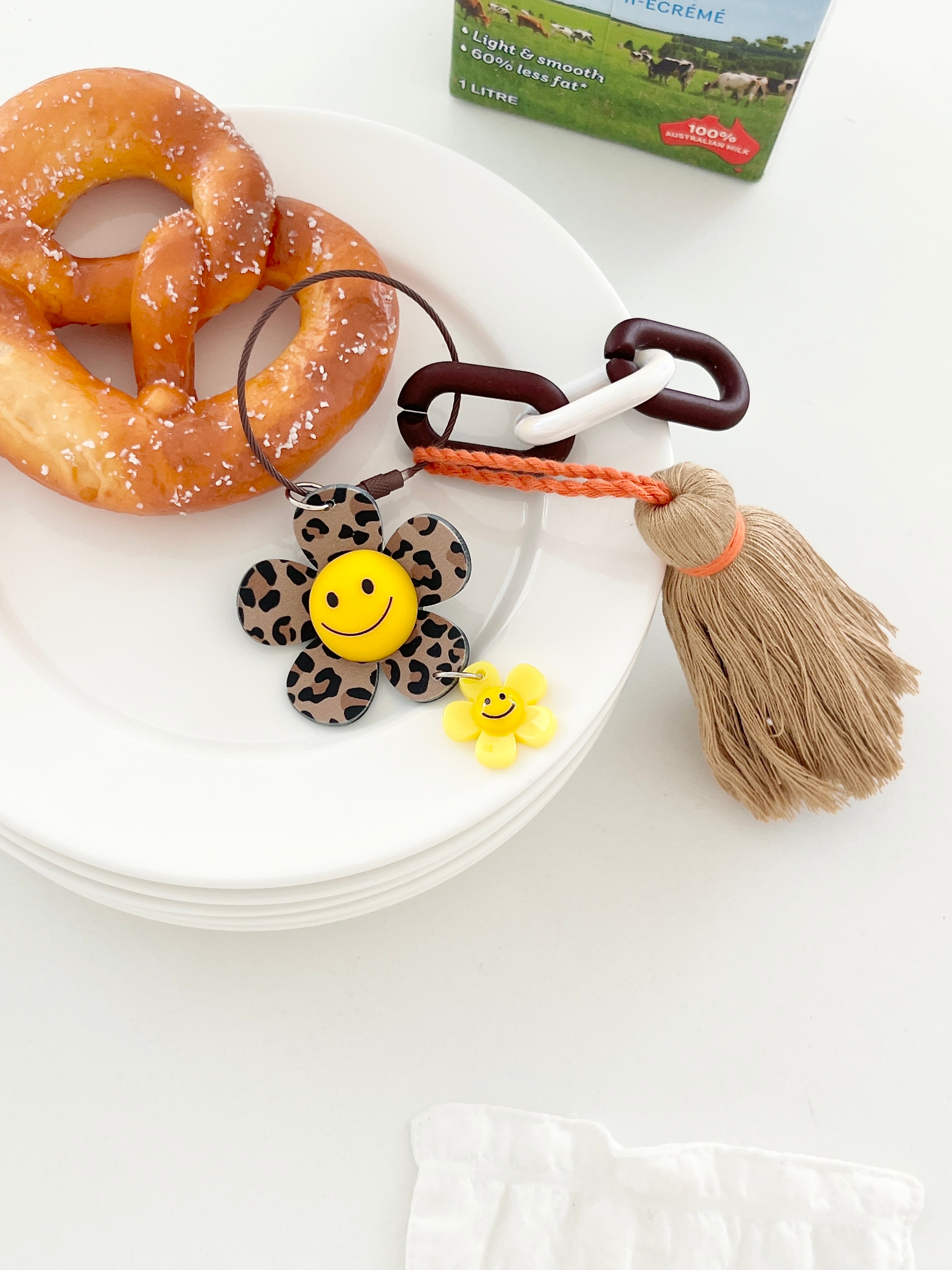 Leopard print flower keychain with a yellow smiley face center, small yellow flower charm, and tan tassel attached to chunky dark brown and white links, placed on a white plate next to a salted pretzel.