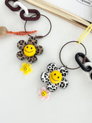 Close-up of two flower-shaped keychains with yellow smiley face centers — one with leopard-print petals and a yellow mini flower charm, the other with black-and-white animal print petals and a pink mini flower charm — showing detailed texture and design of the charms and links.
