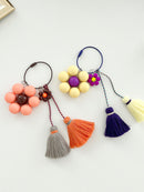 Two keychains are displayed on a white background. The first keychain has a peach flower with a brown center and tassels in gray and orange. The second keychain has a yellow flower with a purple center and tassels in purple and yellow. 