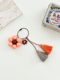 A keychain is displayed on a white background with a peach flower with a brown center and tassels in gray and orange. 
