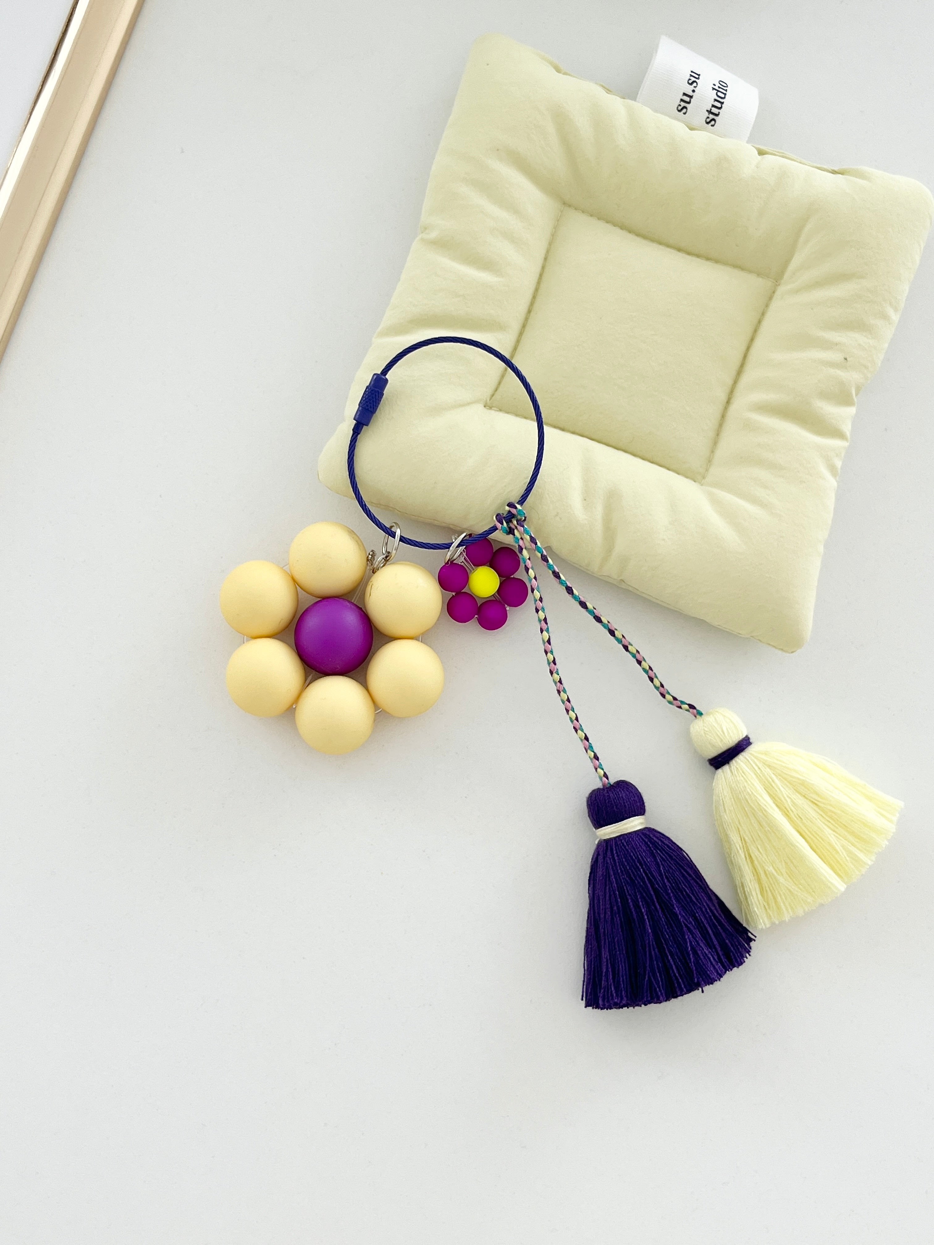 A keychain is displayed on a white background with a yellow flower with a purple center and tassels in purple and yellow. 