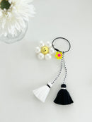 A keychains are displayed on a white background with a white pearl-like flower with a yellow smiley face center and tassels in black and white.