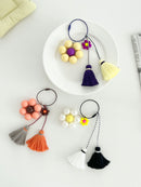 Three keychains are displayed on a white background. The first keychain has a peach flower with a brown center and tassels in gray and orange. The second keychain has a yellow flower with a purple center and tassels in purple and yellow displayed on a white plate. The third keychain has a white pearl-like flower with a yellow smiley face center and tassels in black and white.