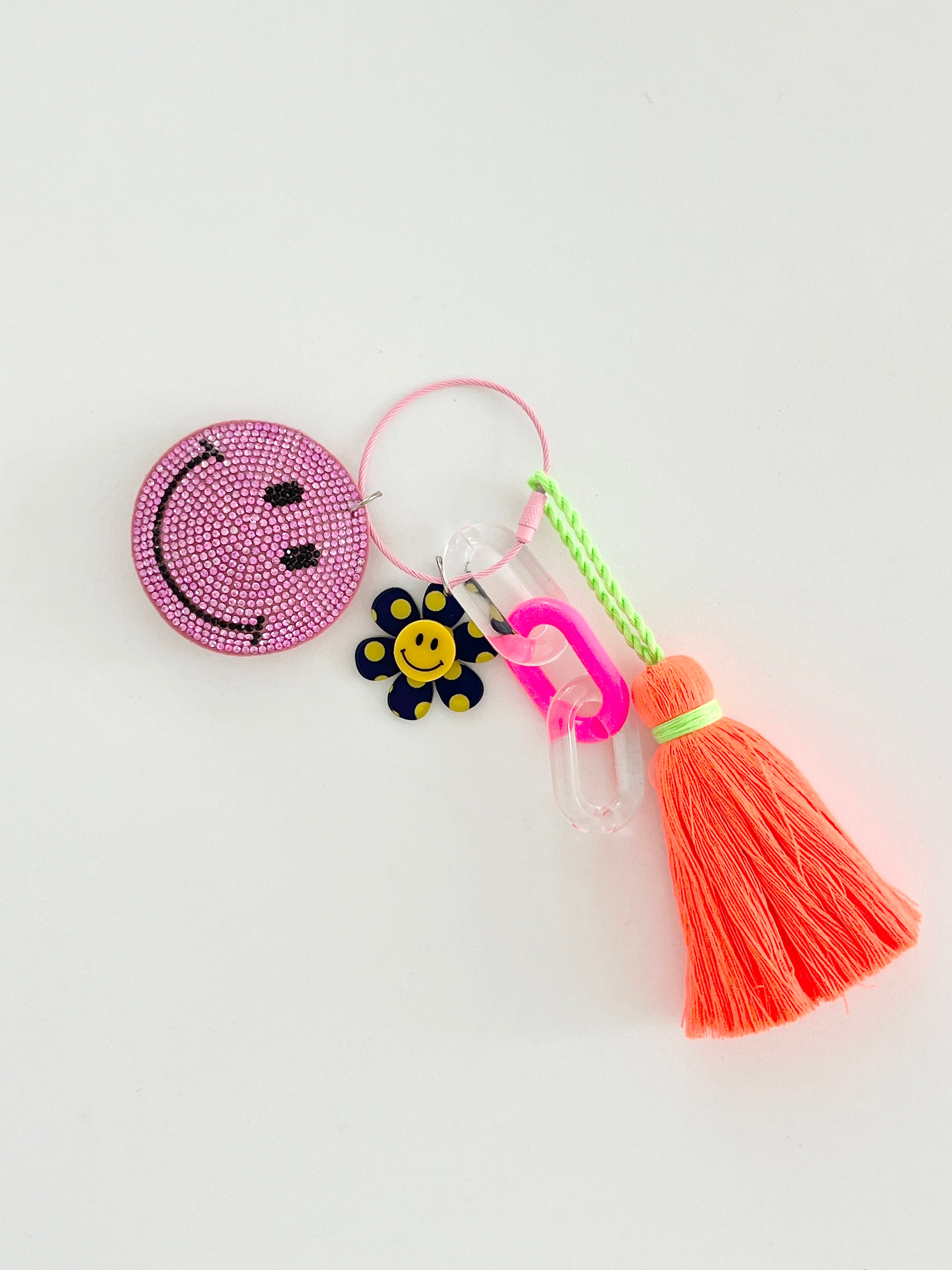 Colorful smiley face keychain with pink rhinestone-covered charms, playful flower accents, and neon orange tassels, displayed on white surface.