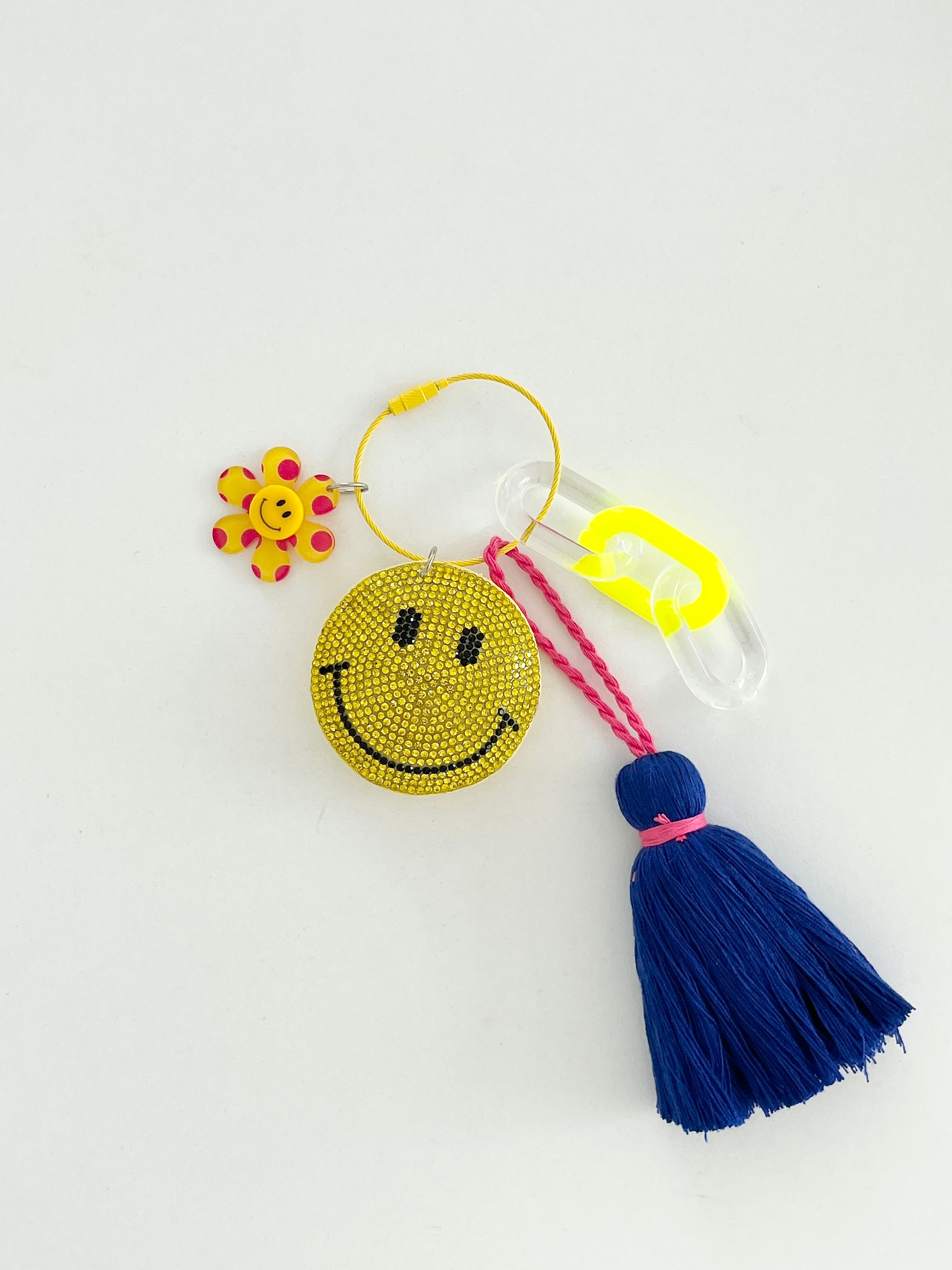 Colorful smiley face keychain with yellow rhinestone-covered charms, playful flower accents, and blue tassels, displayed on white surface.