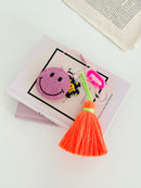 Colorful smiley face keychain with pink rhinestone-covered charms, playful flower accents, and neon orange tassels, displayed on a Dior book with while surface.