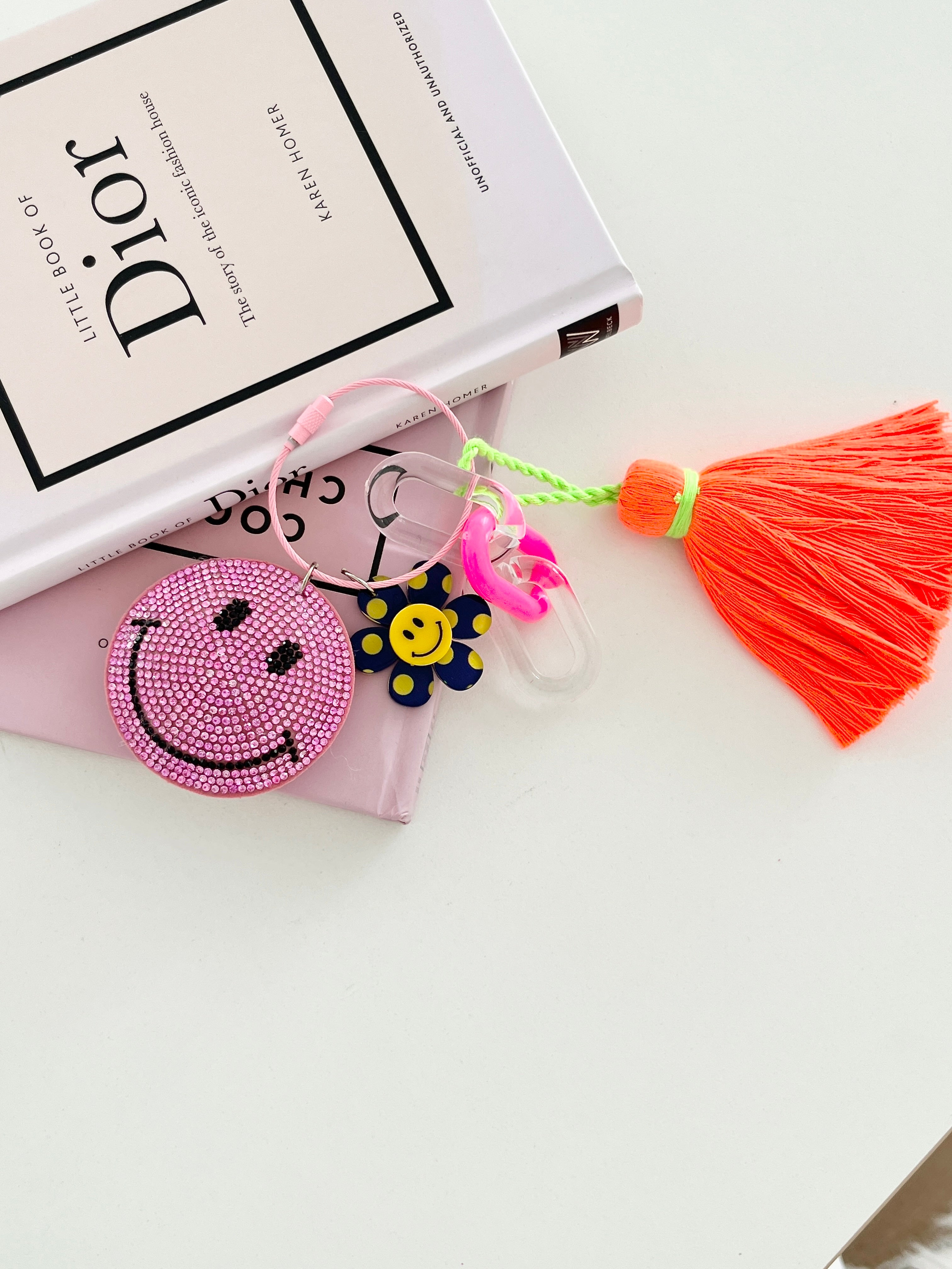 Colorful smiley face keychain with pink rhinestone-covered charms, playful flower accents, and orange tassels, displayed on white surface next to Dior book.