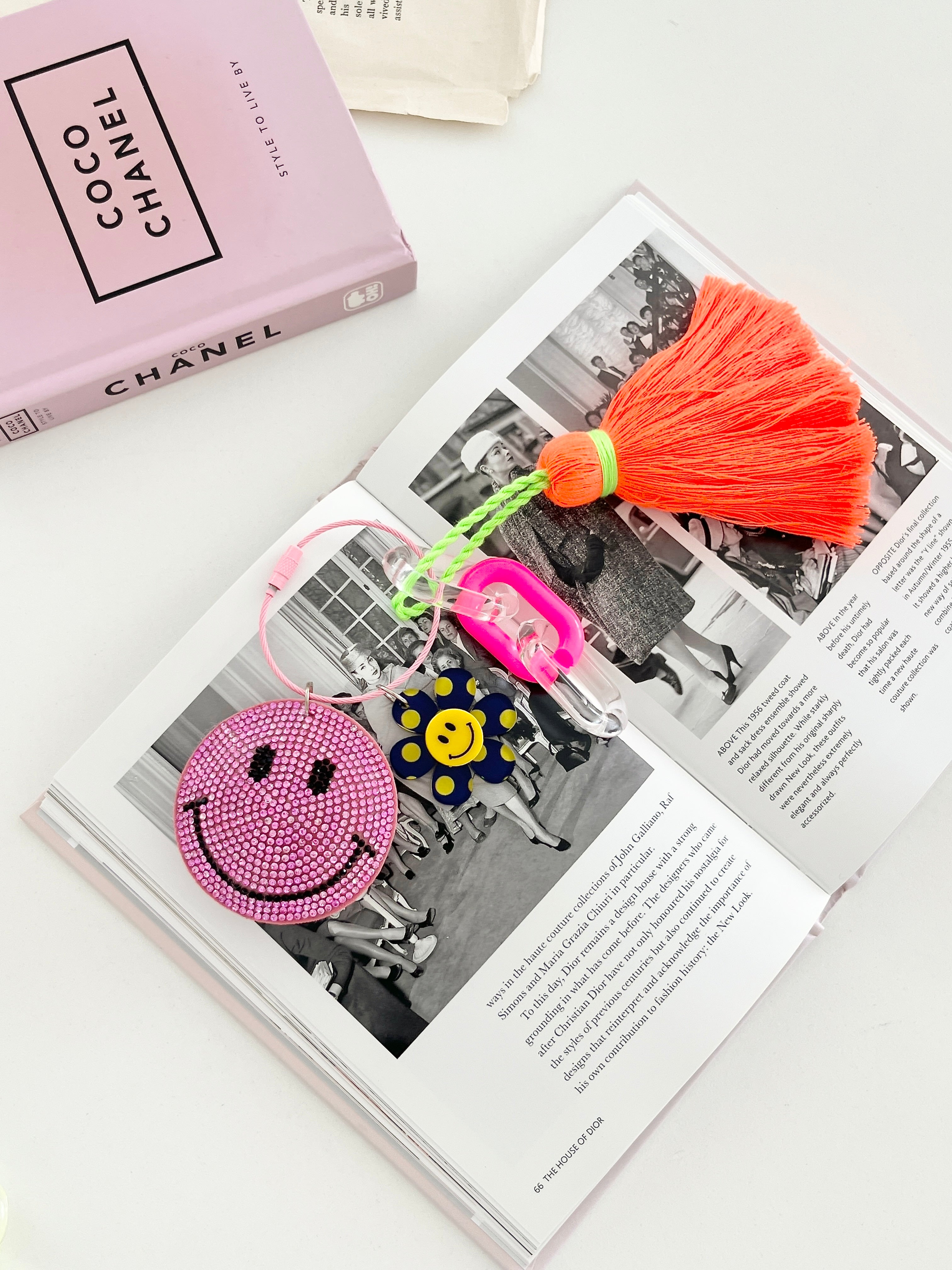 Colorful smiley face keychain with pink rhinestone-covered charms, playful flower accents, and neon orange tassels, displayed on white surface next to pink Chanel book.