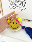Colorful smiley face keychain with yellow rhinestone-covered charms, playful flower accents, and blue tassels, displayed held by a hand on a white surface.