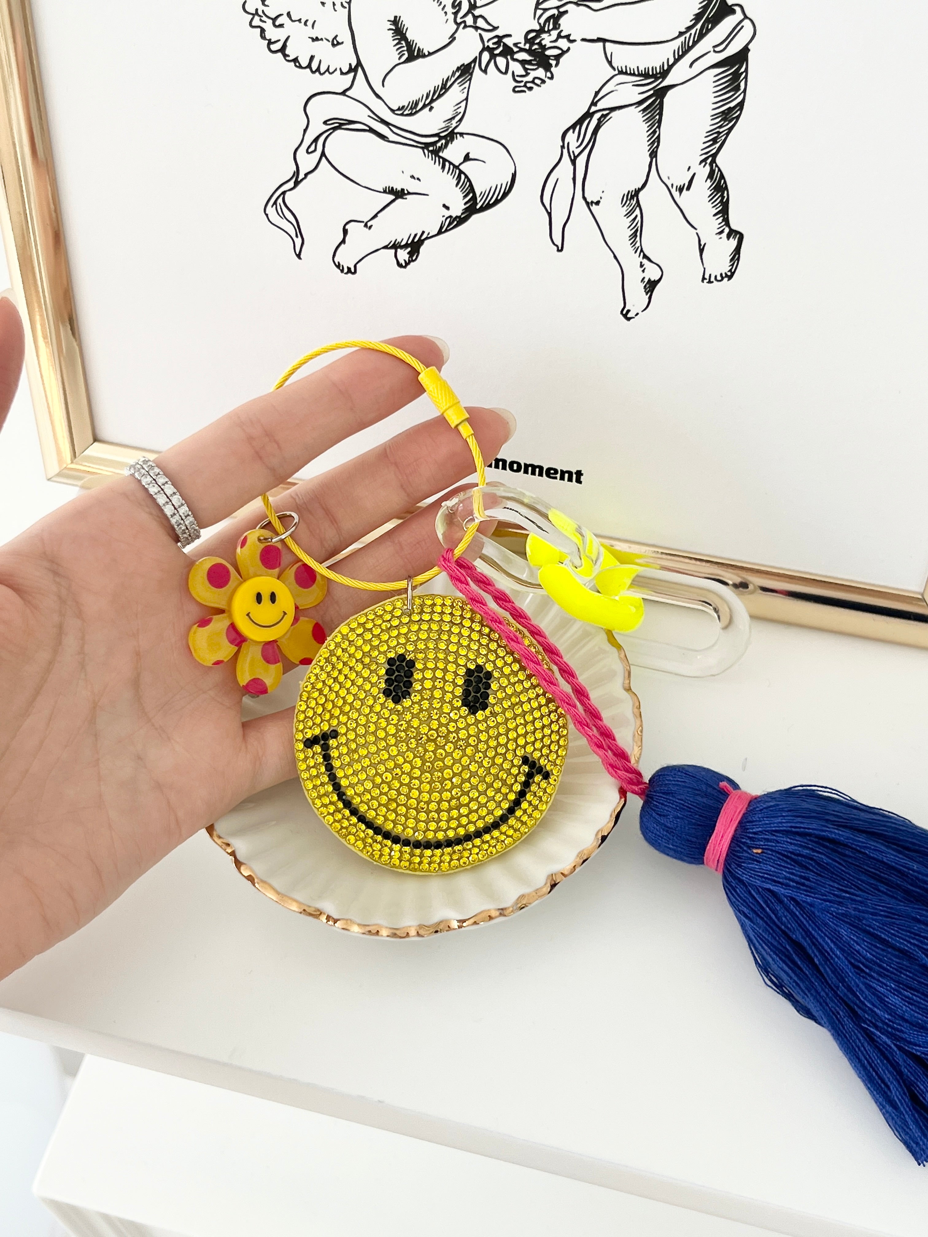 Colorful smiley face keychain with yellow rhinestone-covered charms, playful flower accents, and blue tassels, displayed held by a hand on a white surface.