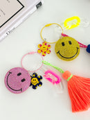 Two colorful smiley face keychains with rhinestone-covered charms, playful flower accents, and bright tassels, displayed on a white surface next to a book.