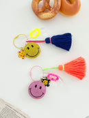 Two colorful smiley face keychains with rhinestone-covered charms, playful flower accents, and  tassels, displayed on white surface with a pretzel and donut in the background.