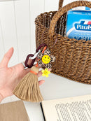 Leopard print flower keychain with a yellow smiley face center, small yellow flower charm, and a tan tassel hanging from chunky dark brown and white links. The keychain is being held in front of a wicker basket containing a milk carton